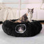 Two cats in a fluffy black pet bed on a light-colored couch.
